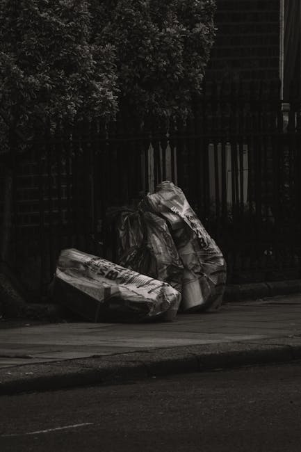 A person's hand and forearm are visible, holding a green plastic rubbish bag tightly by its twisted top, which is gathered and knotted. The bag appears to be filled with waste, as it is slightly bulgi