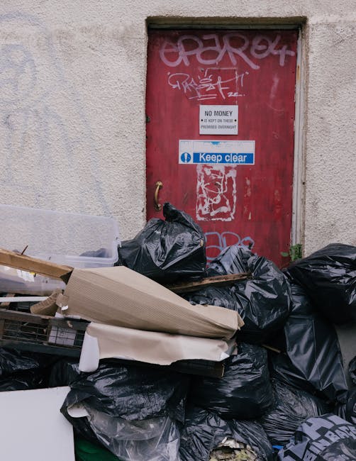 A collection of black plastic rubbish bags stacked in front of a red metal door with graffiti and stickers, situated on an exterior wall of a building with weathered, light-colored plaster. The debris
