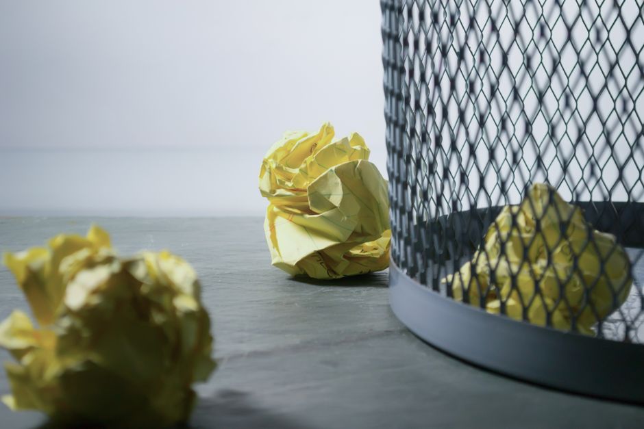 A collection of black plastic rubbish bags stacked in front of a red metal door with graffiti and stickers, situated on an exterior wall of a building with weathered, light-colored plaster. The debris